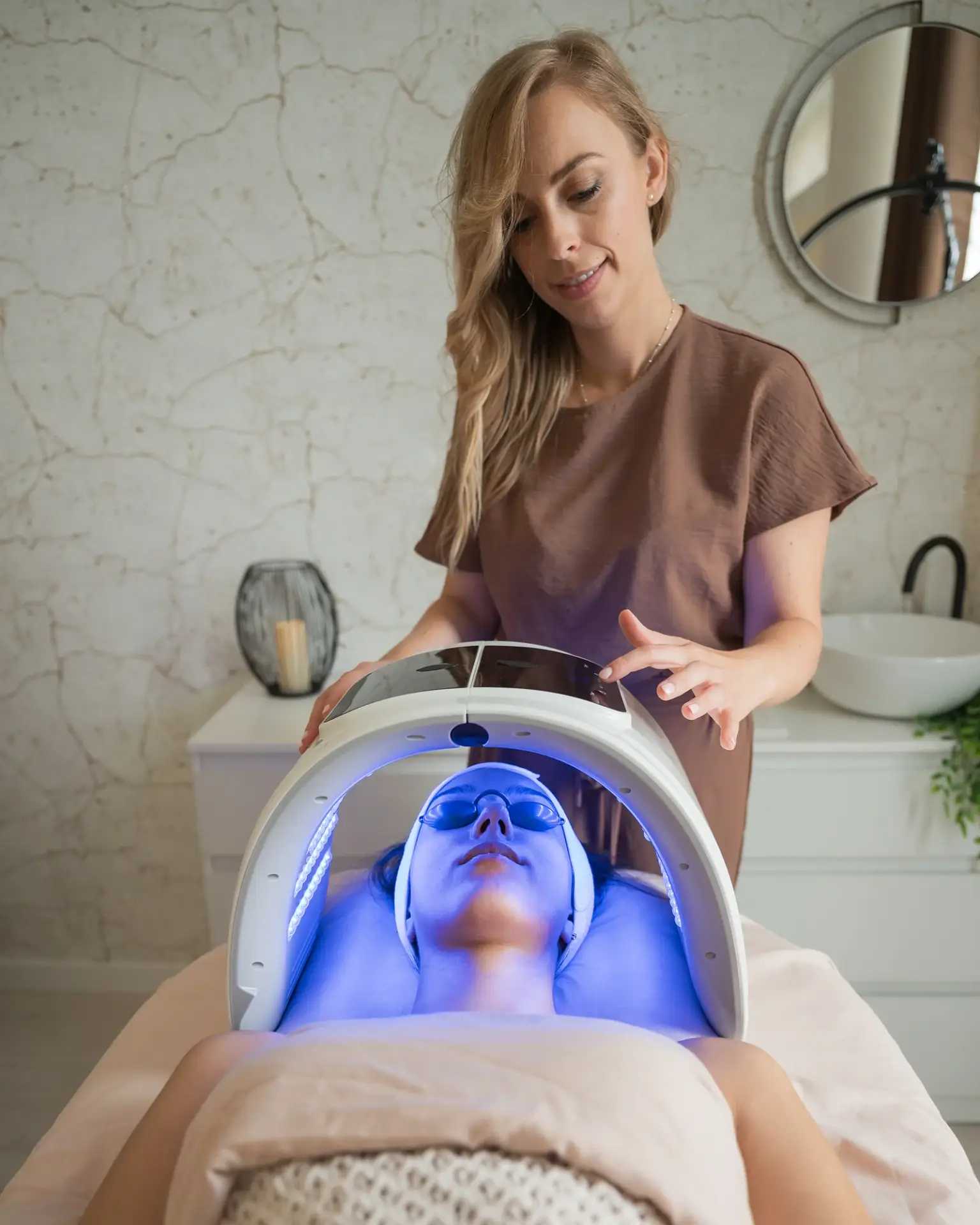 Esthetician guiding a patient through an LED light facial treatment in a bright med spa treatment room