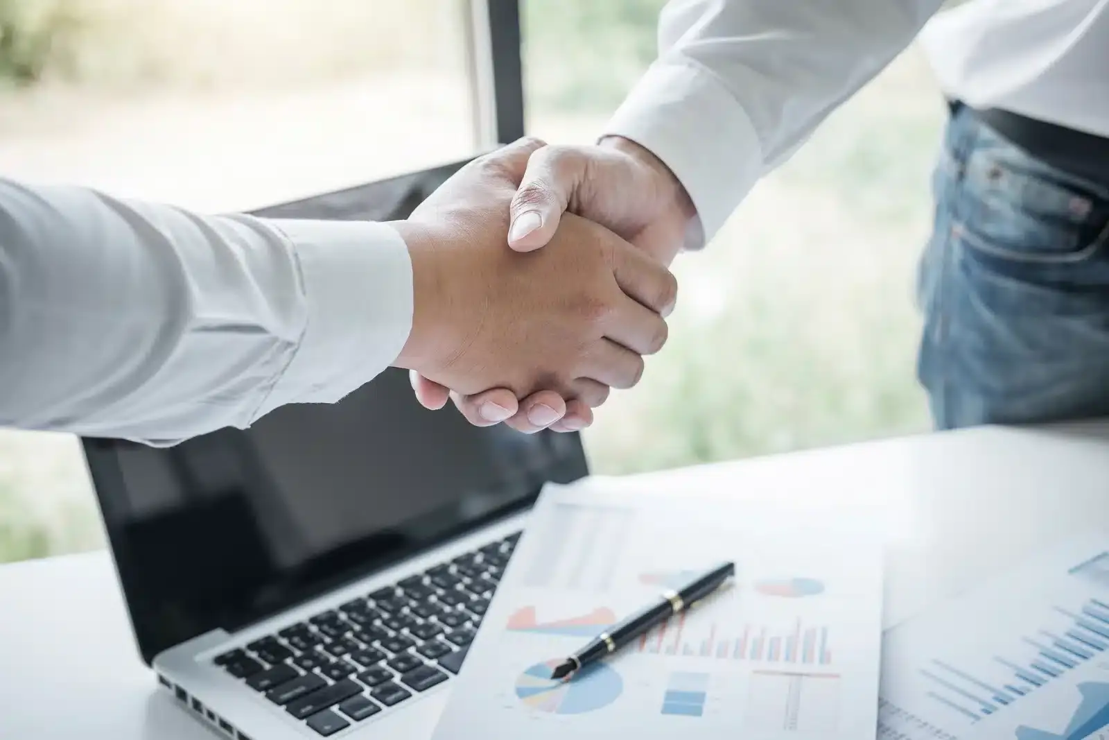 Two professionals shaking hands over a desk with charts and a laptop, illustrating a transparent plastic surgery SEO agreement.